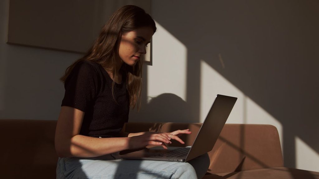 Serious young female in casual outfit sitting on cozy couch and typing on contemporary netbook in shadow light